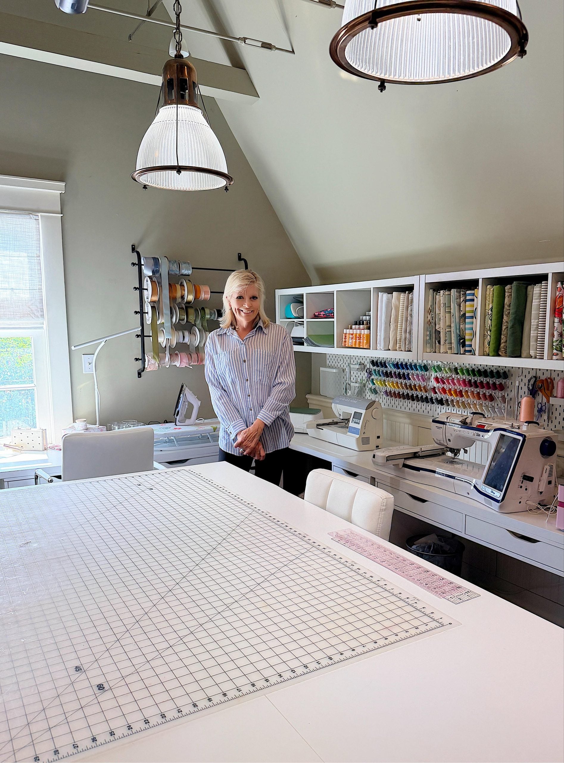 A woman stands in a well-organized sewing room with fabric, ribbons, sewing machines, and tools neatly arranged on shelves and a large cutting table in the foreground.
