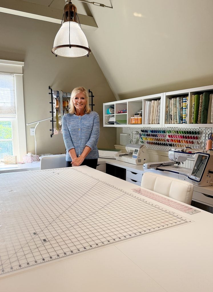 A woman stands in a bright sewing room, surrounded by fabric, thread, and sewing machines, with a large cutting mat on the table in front of her.