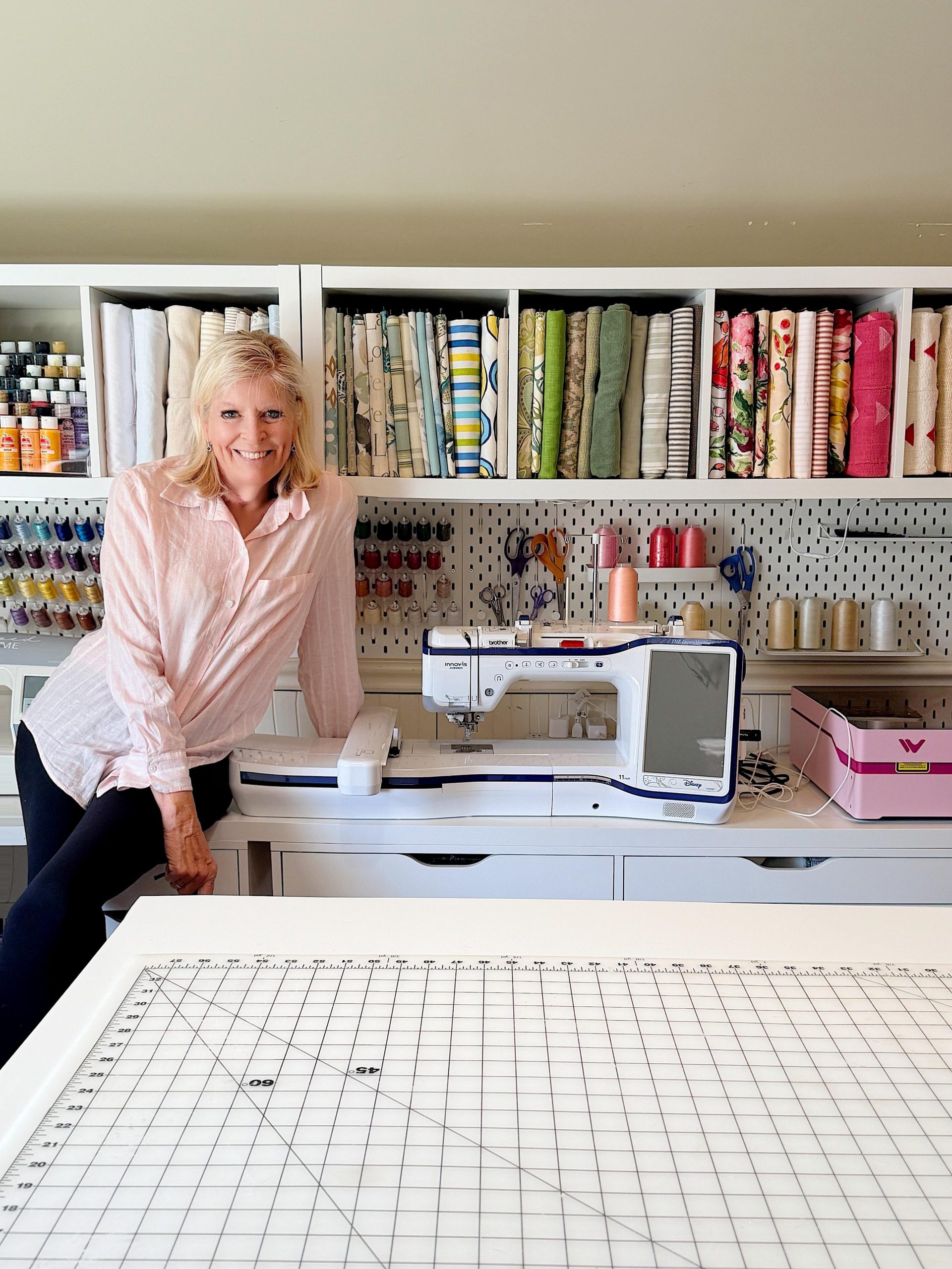 A woman stands next to a sewing machine in a craft room with shelves of fabric, thread, and supplies in the background.