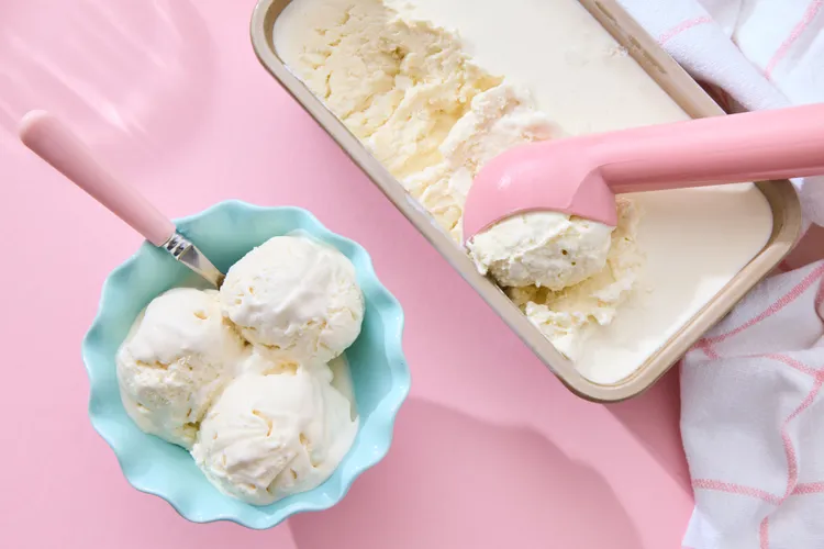 A bowl with three scoops of vanilla ice cream sits next to a rectangular container of ice cream and a pink ice cream scoop on a pink surface.