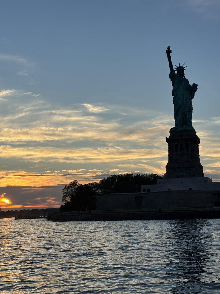 The Statue of Liberty stands silhouetted against a sunset sky, with water in the foreground and trees at the statue’s base.