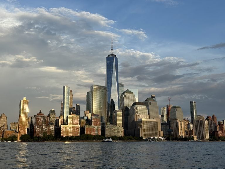 A city skyline with tall modern buildings, including the One World Trade Center, viewed from across a body of water under a partly cloudy sky.