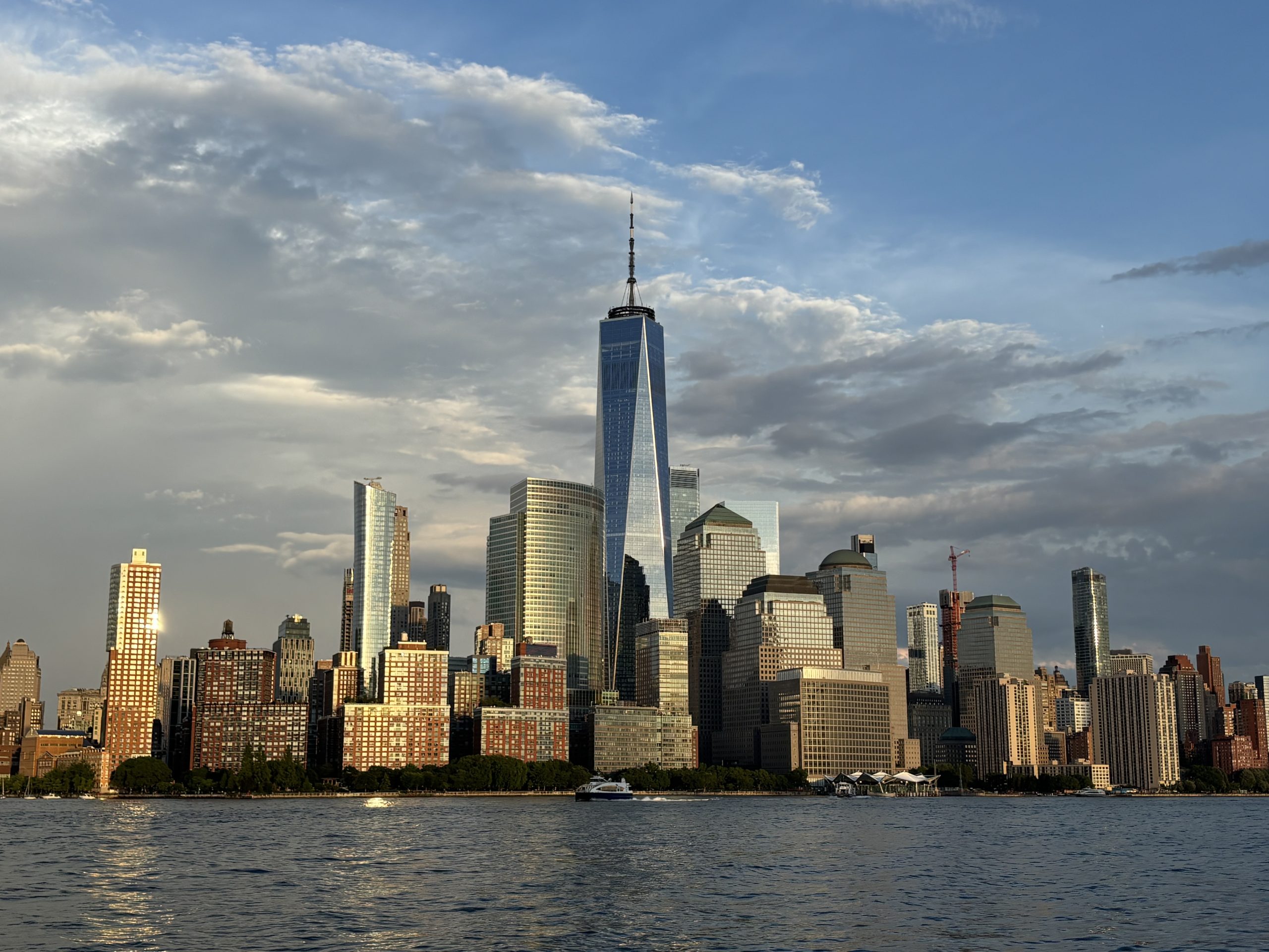 A city skyline with tall modern buildings, including the One World Trade Center, viewed from across a body of water under a partly cloudy sky.