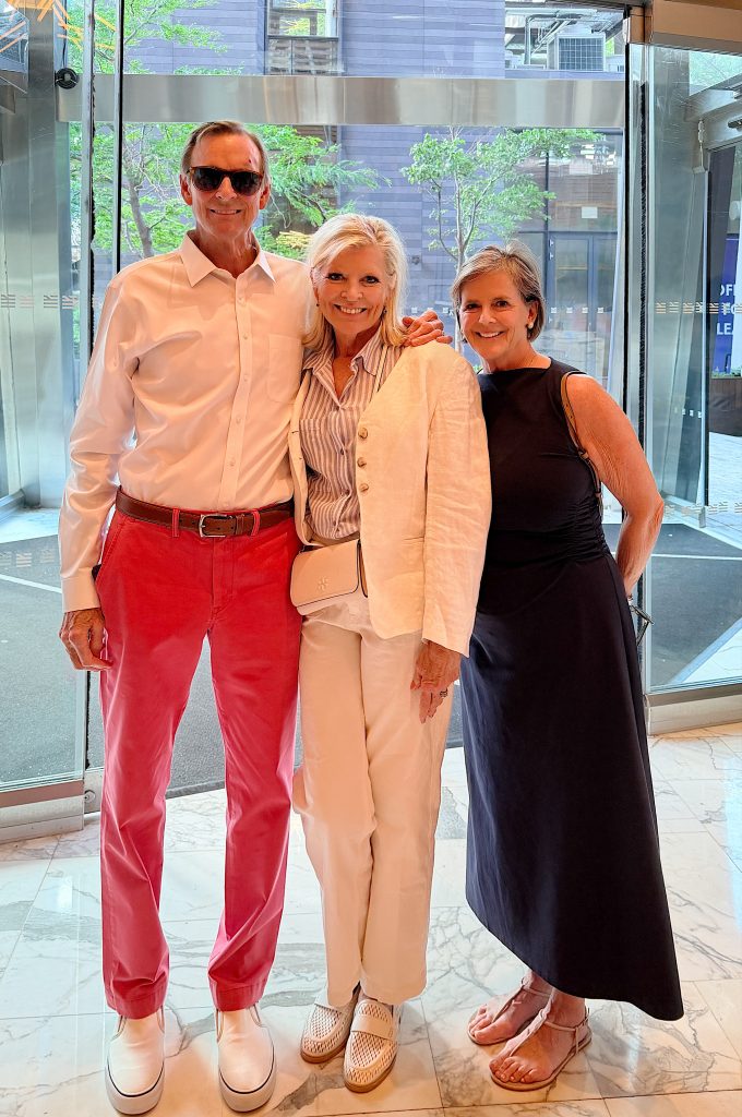 Three adults stand together in a lobby with glass doors and trees outside. One man wears a white shirt and red pants; two women wear light-colored and black outfits, smiling at the camera.