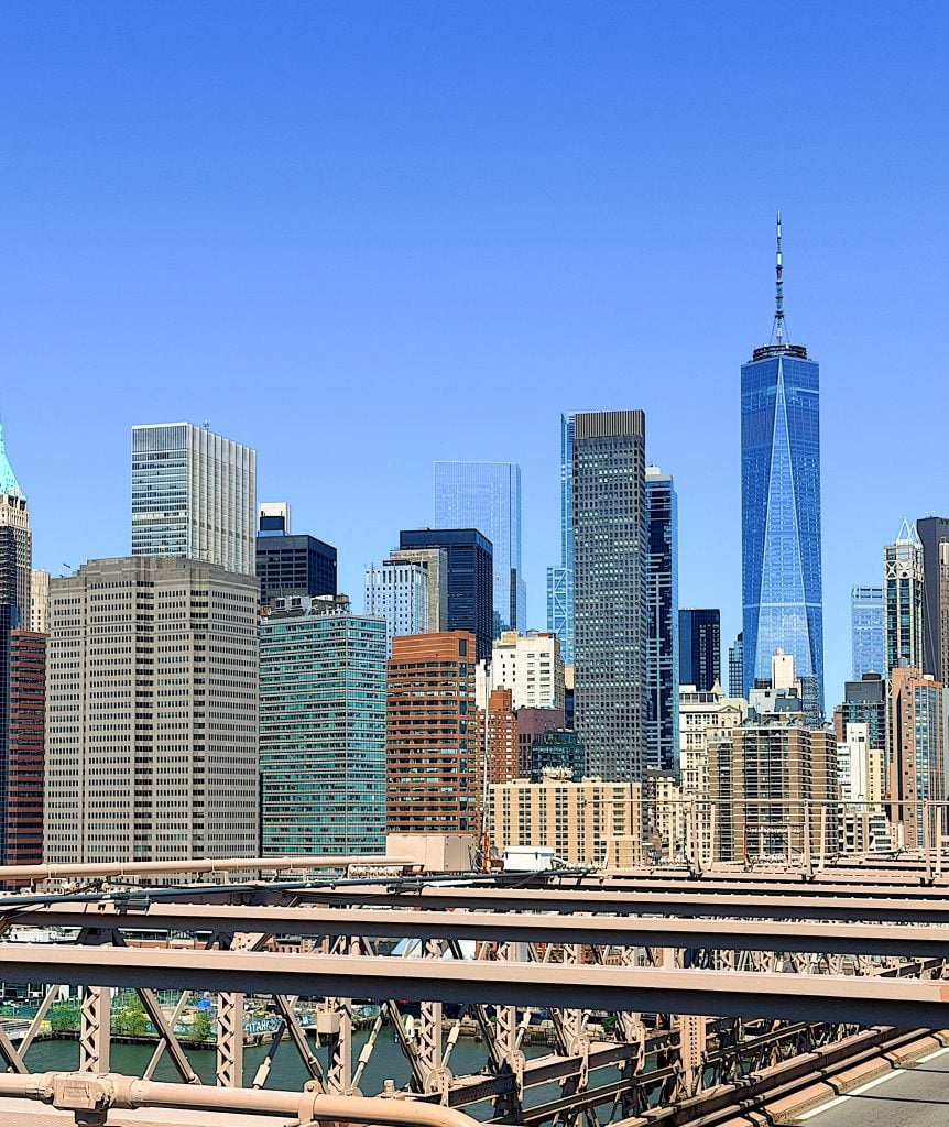 A city skyline with tall modern buildings under a clear blue sky, seen from a bridge with metal beams in the foreground.