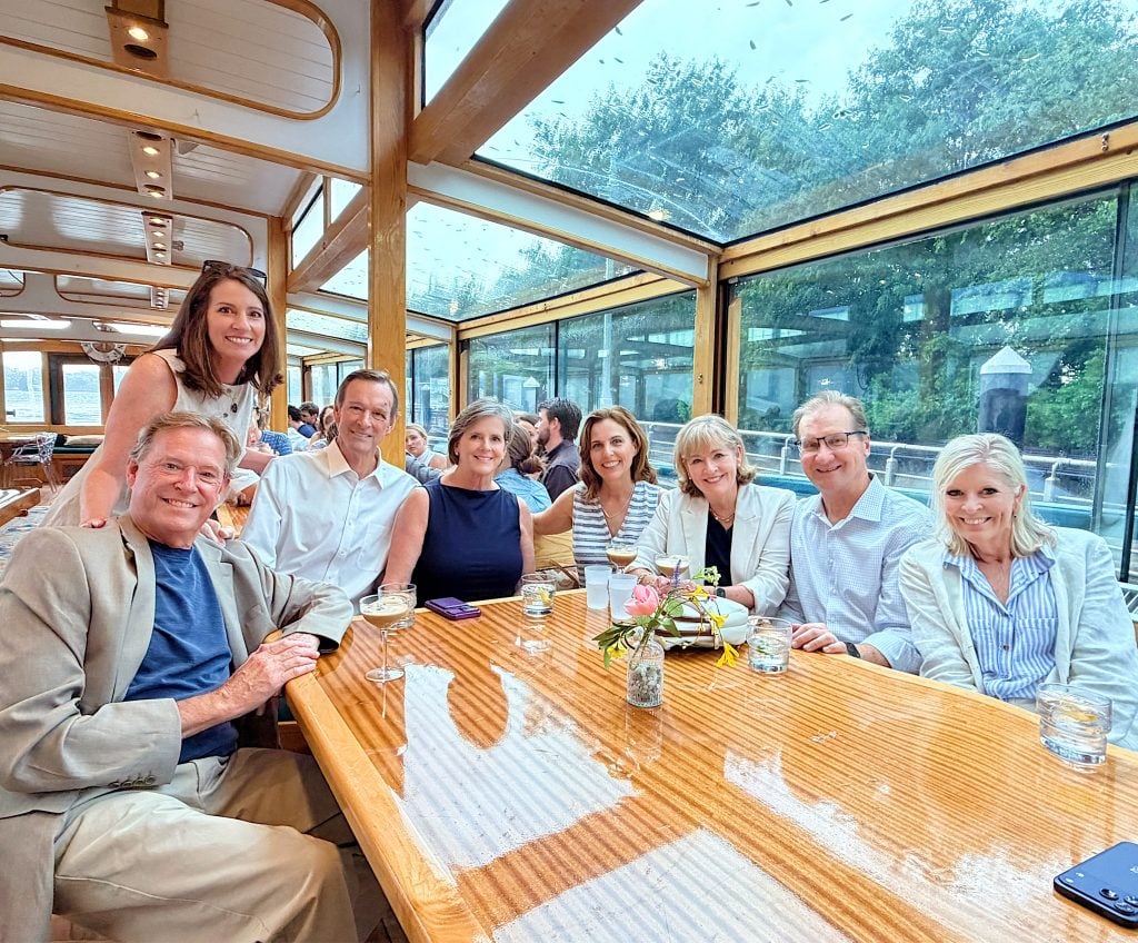 Eight adults sit around a wooden table on a boat with large glass windows, smiling at the camera. Drinks and a small floral centerpiece are on the table. Trees are visible outside.
