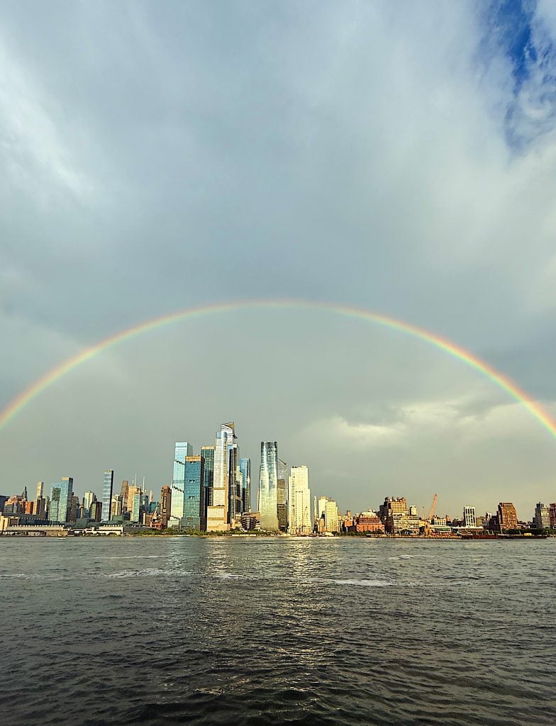A rainbow arches over a city skyline with tall modern buildings next to a river under a partly cloudy sky.