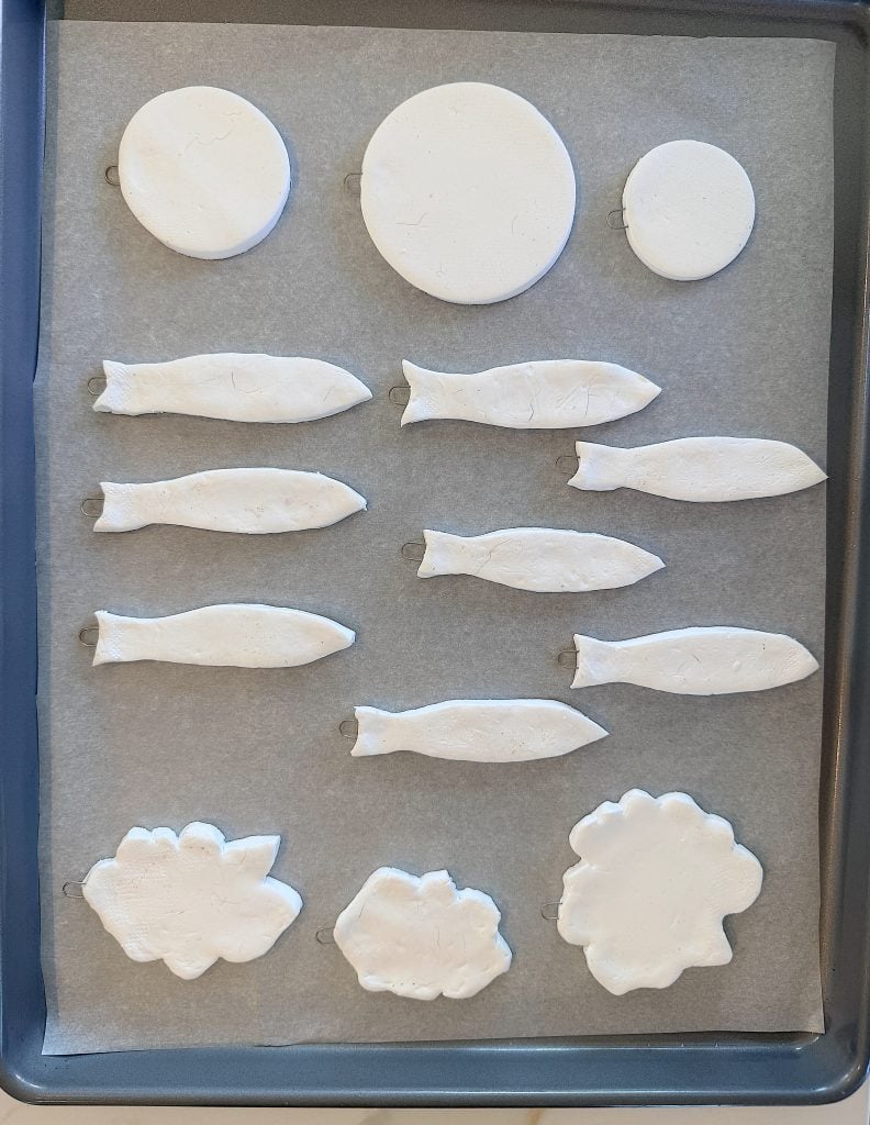 A baking tray with parchment paper holds unbaked clay shapes: two circles, eight fish, and three irregular blobs arranged in rows.