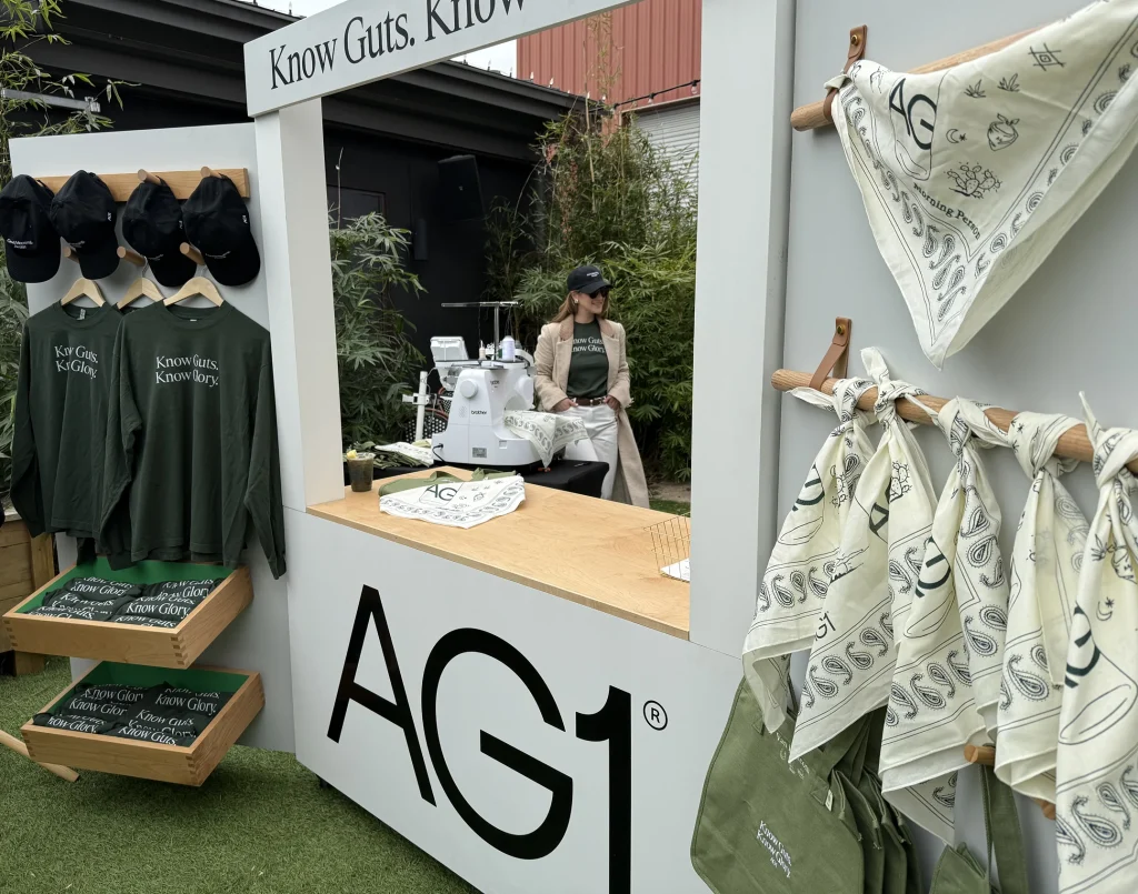 AG1 promotional booth featuring green shirts, hats, and white bandanas on display, with a person seated behind a sewing machine in the background.