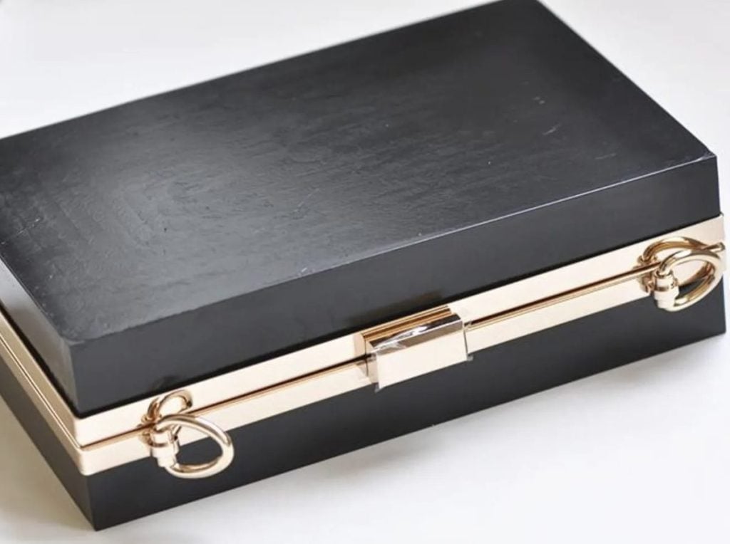 A rectangular black box with gold metal trim and two circular gold handles on each end, placed on a white surface.