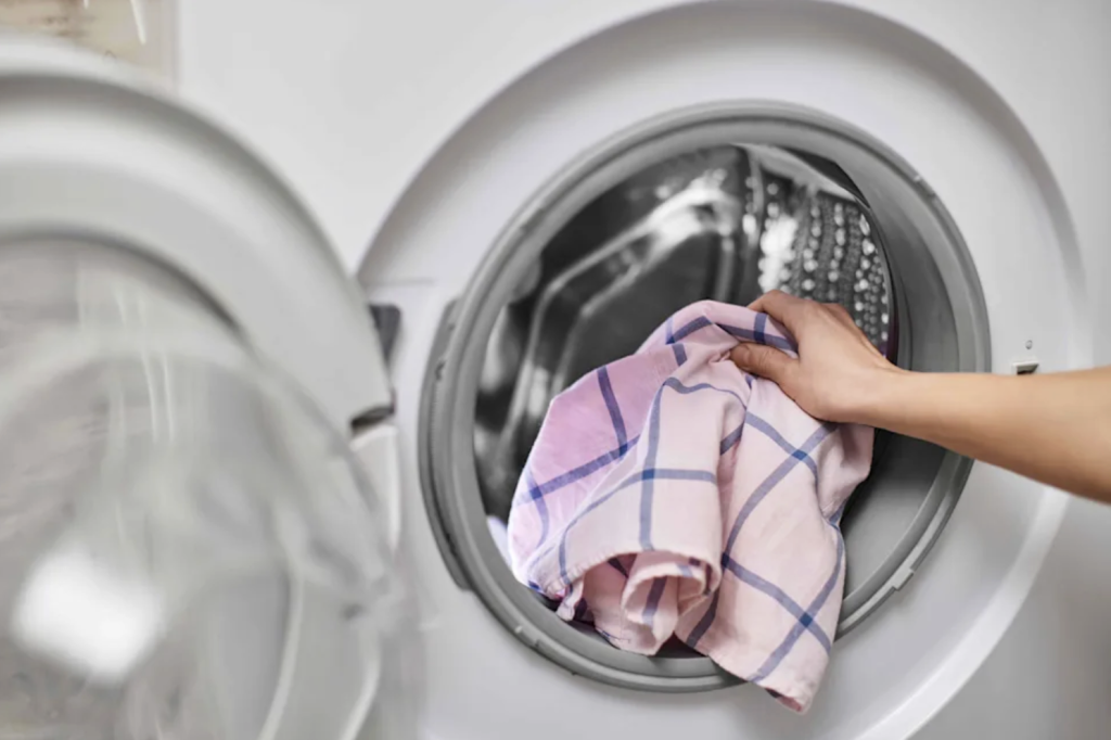 A hand places a pink and blue plaid shirt into the open door of a front-loading washing machine.