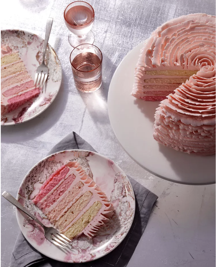 A pink ombre layer cake with decorative frosting sits on a cake stand. Two slices are served on patterned plates with forks, next to pink glasses filled with water.