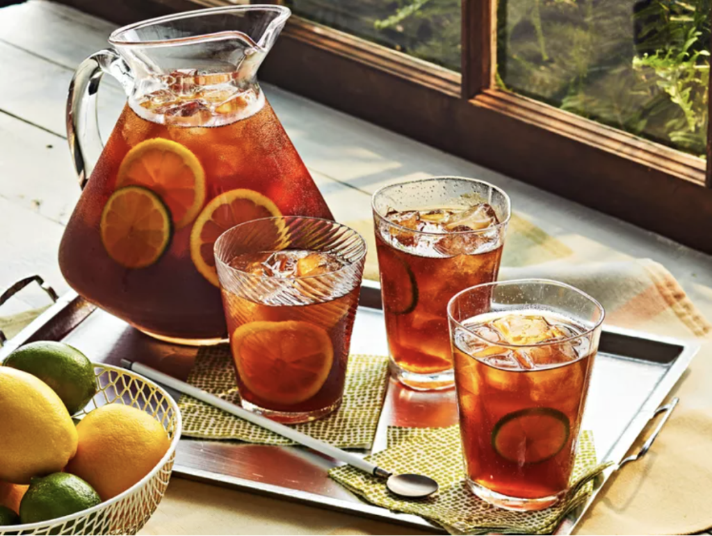 A pitcher and three glasses of iced tea with lemon and lime slices sit on a tray near a bowl of citrus fruit and a spoon by a sunlit window.