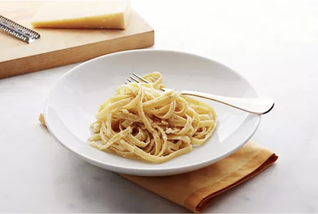 A white plate of fettuccine pasta with a fork, set on a napkin, with a wedge of cheese and grater in the background on a cutting board.