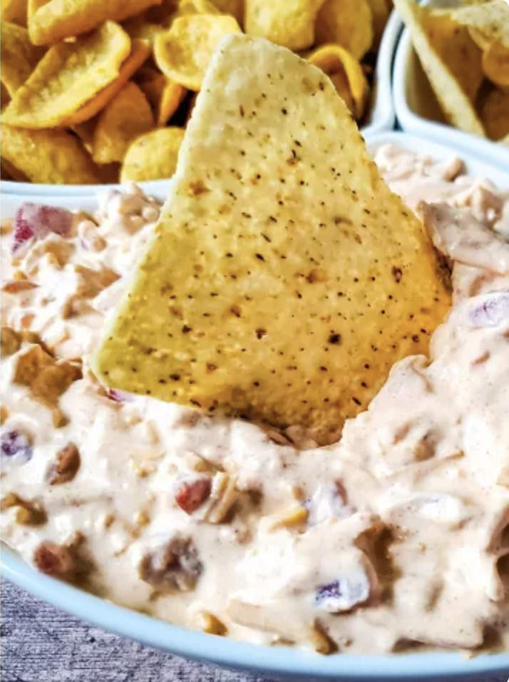 A tortilla chip dipped into a creamy, chunky salsa dip in a white bowl, with corn chips visible in the background.