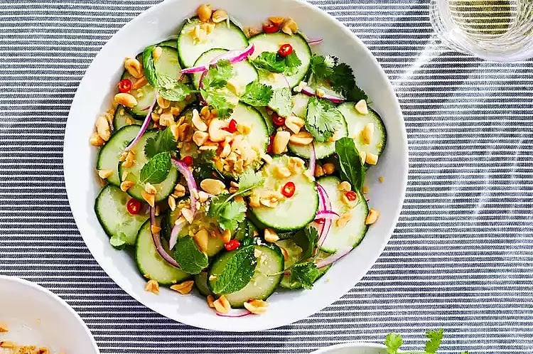 A bowl of cucumber salad with sliced red onions, fresh herbs, chopped peanuts, and red chili on a striped tablecloth.