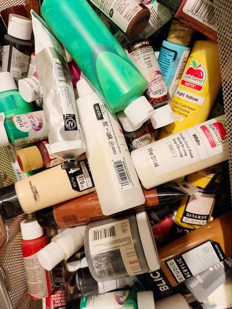 Assorted tubes and bottles of acrylic paint in various colors scattered inside a mesh container.