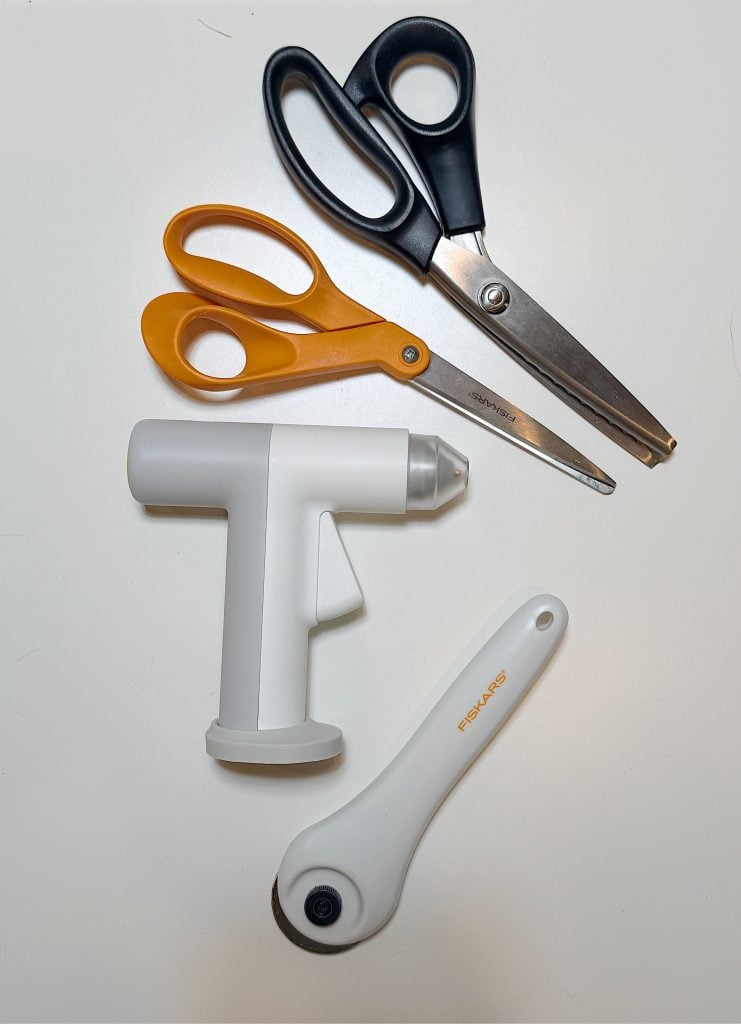 Two pairs of scissors, a white handheld sewing tool, and a white rotary cutter are arranged on a white surface.