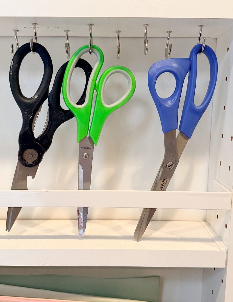 Three pairs of scissors—black, green, and blue—hang by their handles on hooks above a white shelf with a pegboard to the side.