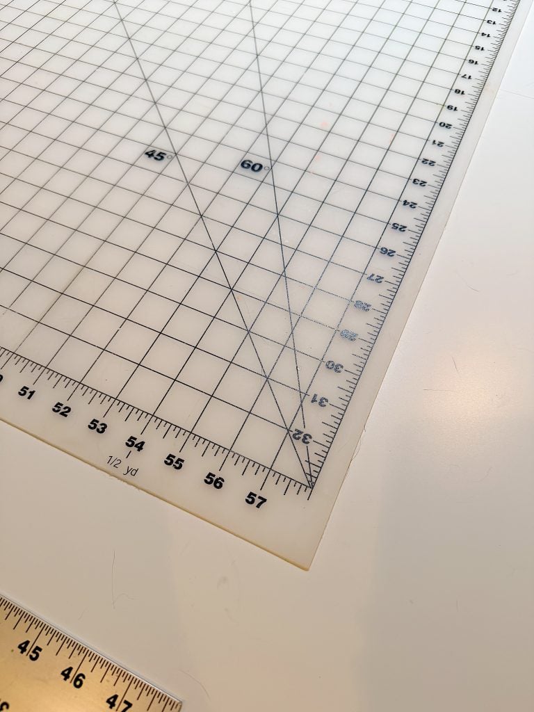 A close-up of a gridded cutting mat with marked measurement lines and angles, placed on a white table. A ruler is partially visible in the corner.