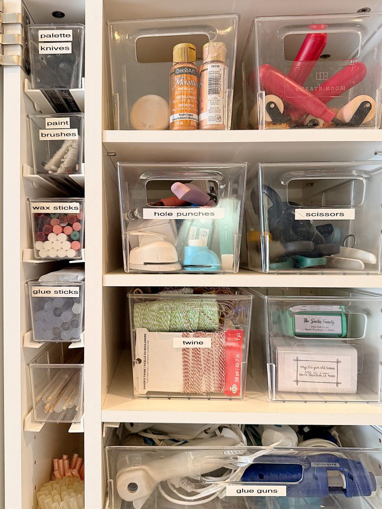 Clear plastic bins labeled and organized on shelves contain various craft supplies, including glue guns, twine, scissors, hole punches, paint brushes, wax sticks, and palette knives.