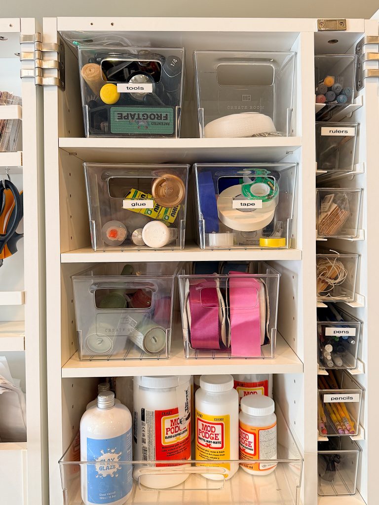 Organized craft supplies on white shelves, including labeled bins with tools, glue, tape, ribbon, clay, Mod Podge, and other materials.
