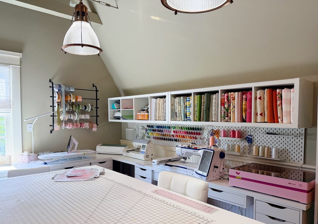 A well-organized sewing and craft room with machines, shelves of fabric, spools of thread, ribbon racks, and a large cutting table under bright lighting.