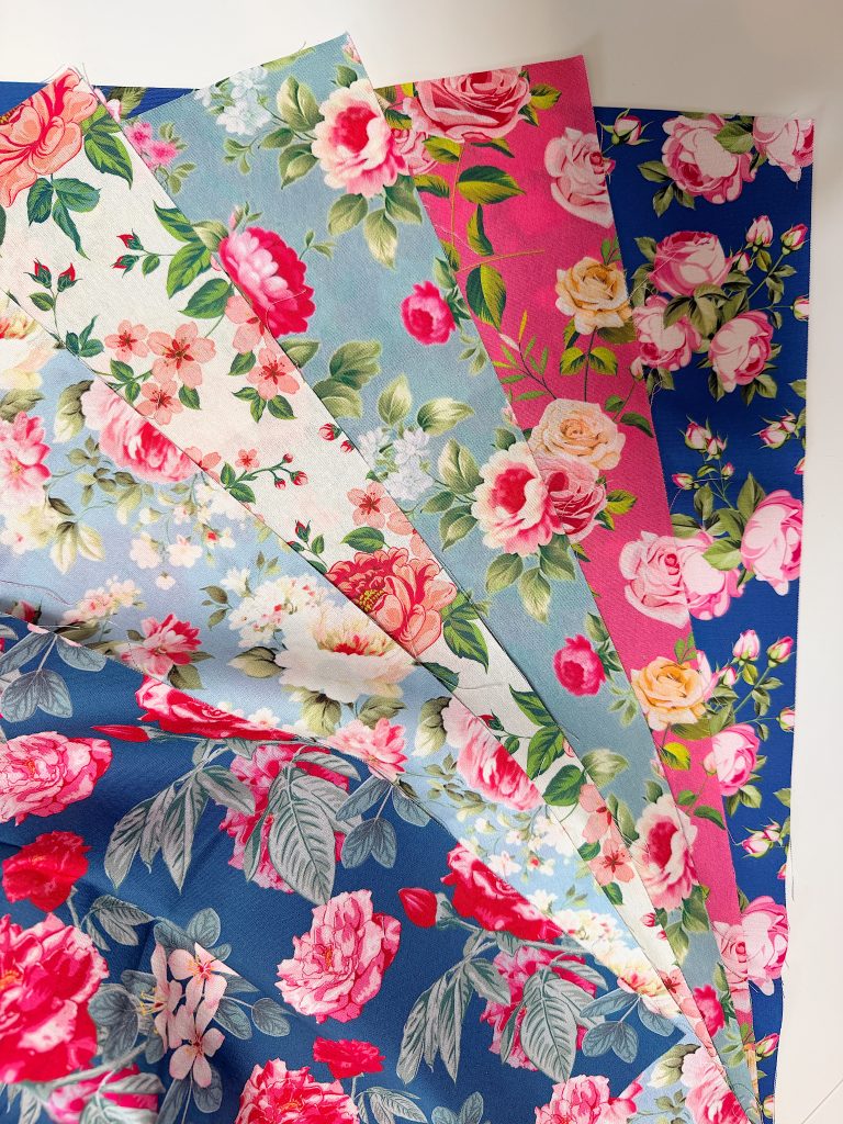 Five sheets of fabric with different floral patterns in shades of pink, blue, and green are fanned out on a flat surface.