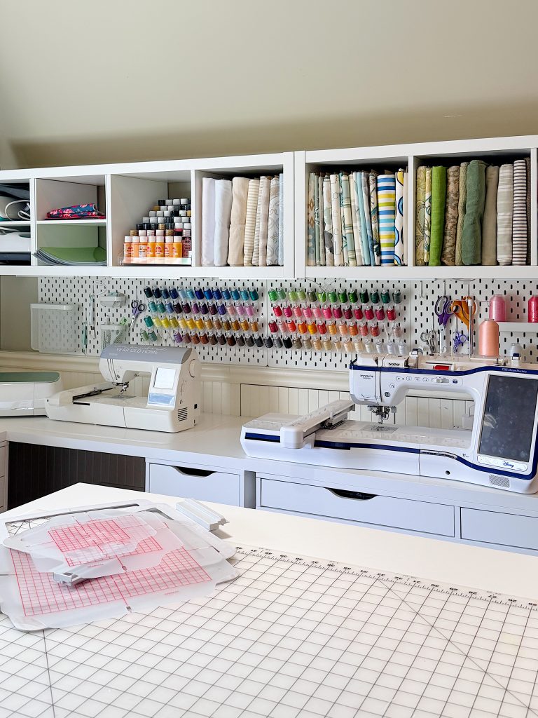 A well-organized sewing room with sewing machines, shelves of fabric and thread, tools, and supplies neatly arranged on and above a white worktable.