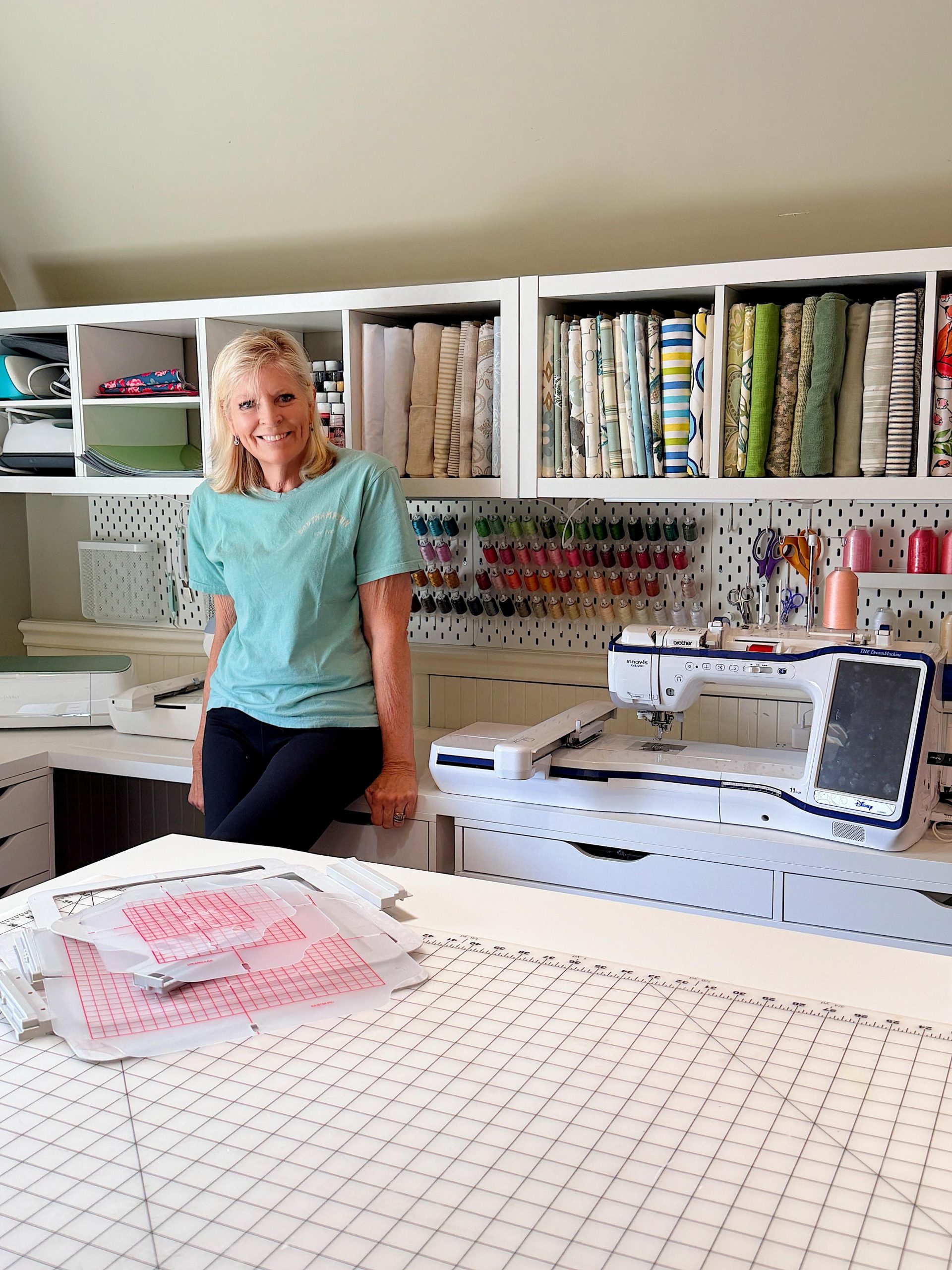 A woman stands in a sewing room with organized fabric, sewing supplies, a sewing machine, and a cutting mat on the table.