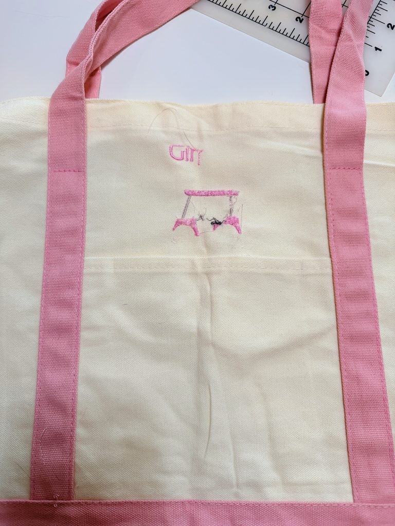 A white tote bag with pink handles and trim, featuring the word "Girl" and a partially embroidered design in pink and black thread on the front pocket. A ruler is visible in the corner.