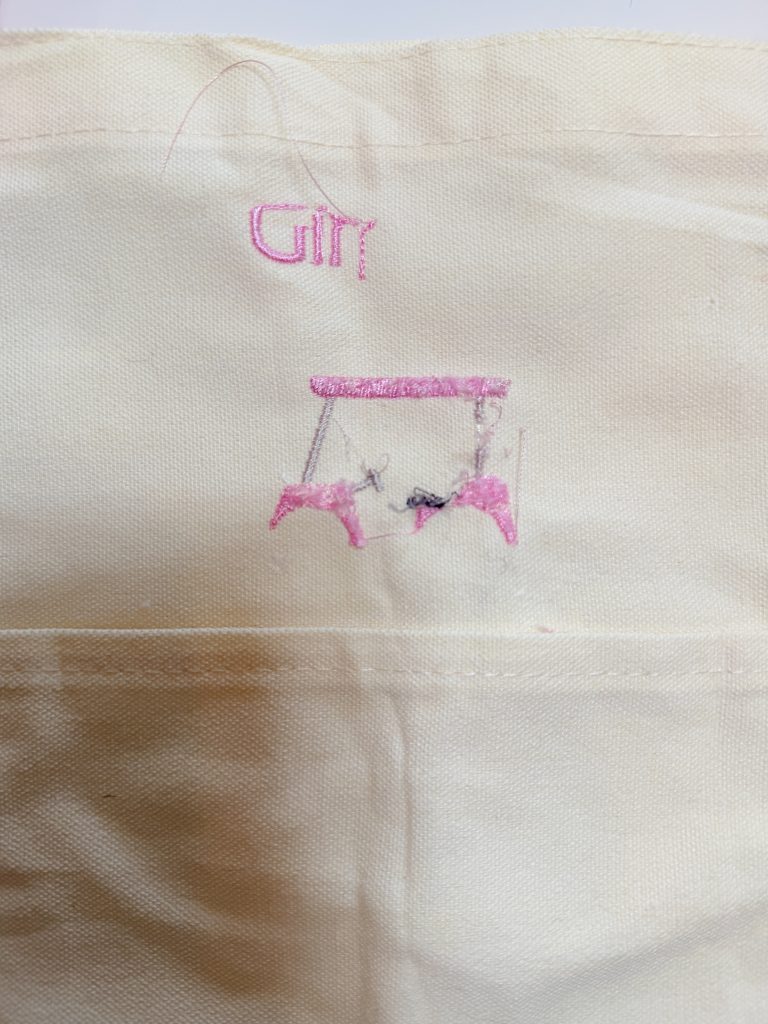 A piece of cream fabric with pink and gray embroidery showing the word "Girl" above two abstract pink shapes and some loose threads.