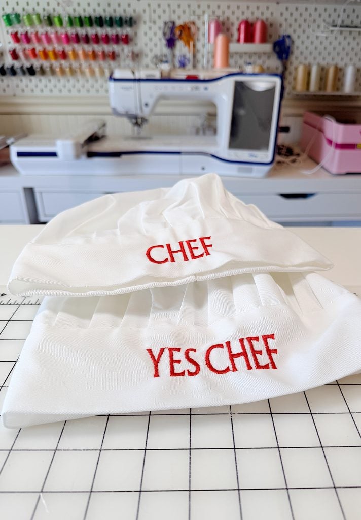 Two white chef hats with red embroidered text, "CHEF" and "YES CHEF," are displayed on a grid-patterned table in a sewing room with supplies and a sewing machine in the background.