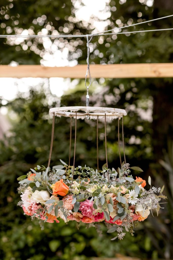 A circular floral arrangement with orange and pink flowers and greenery hangs from wires outdoors against a background of trees and wooden beams.