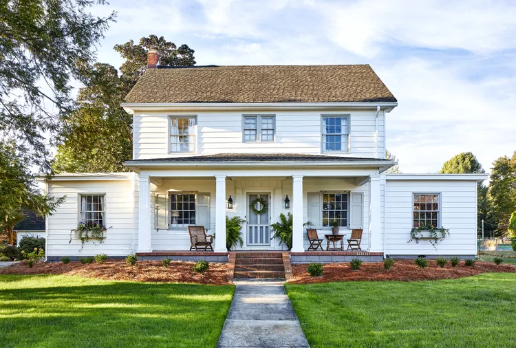 Two-story white house with a covered front porch, two side wings, potted plants, outdoor seating, and a well-kept lawn.