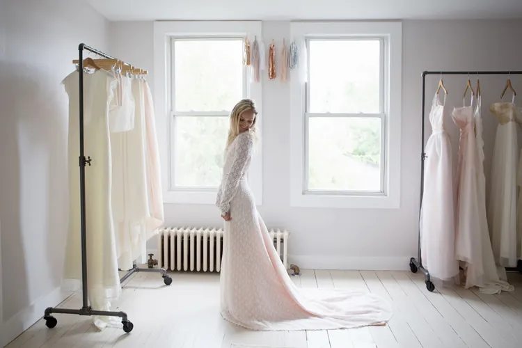 A woman in a long white wedding dress stands in a bright room with racks of dresses and two large windows in the background.
