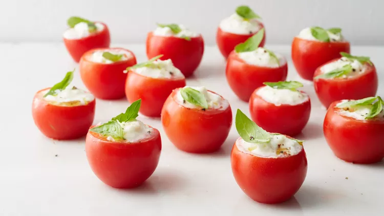 Cherry tomatoes filled with a creamy white mixture, each topped with a fresh basil leaf, arranged on a white surface.