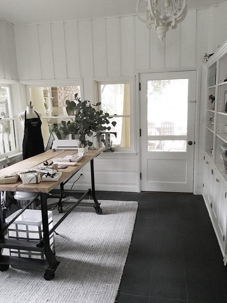 A bright, organized workspace with a wooden desk, shelves, a mannequin, and a large rug, set against white walls and windows letting in natural light.