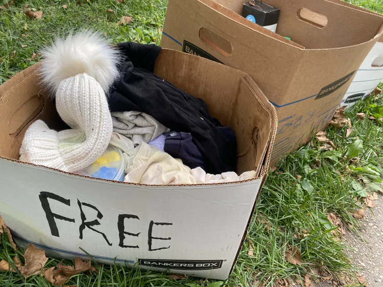 Two cardboard boxes sit on grass; one contains assorted clothing and a white knit hat, with "FREE" handwritten on the front.