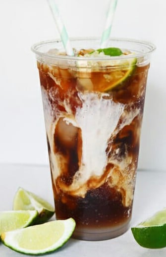 A plastic cup filled with iced coffee or cola, swirled with cream, garnished with lime slices, and two striped straws; lime wedges are placed nearby.