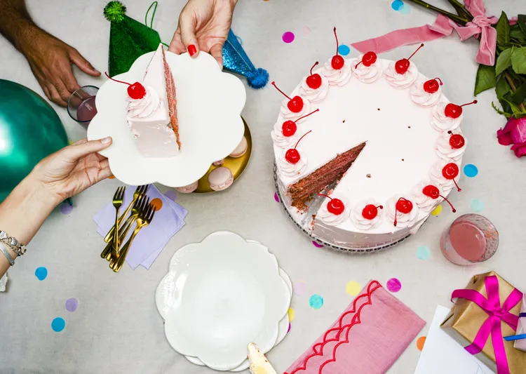 A person serves a slice of cherry-topped cake at a decorated table with party hats, gifts, confetti, flowers, and plates.