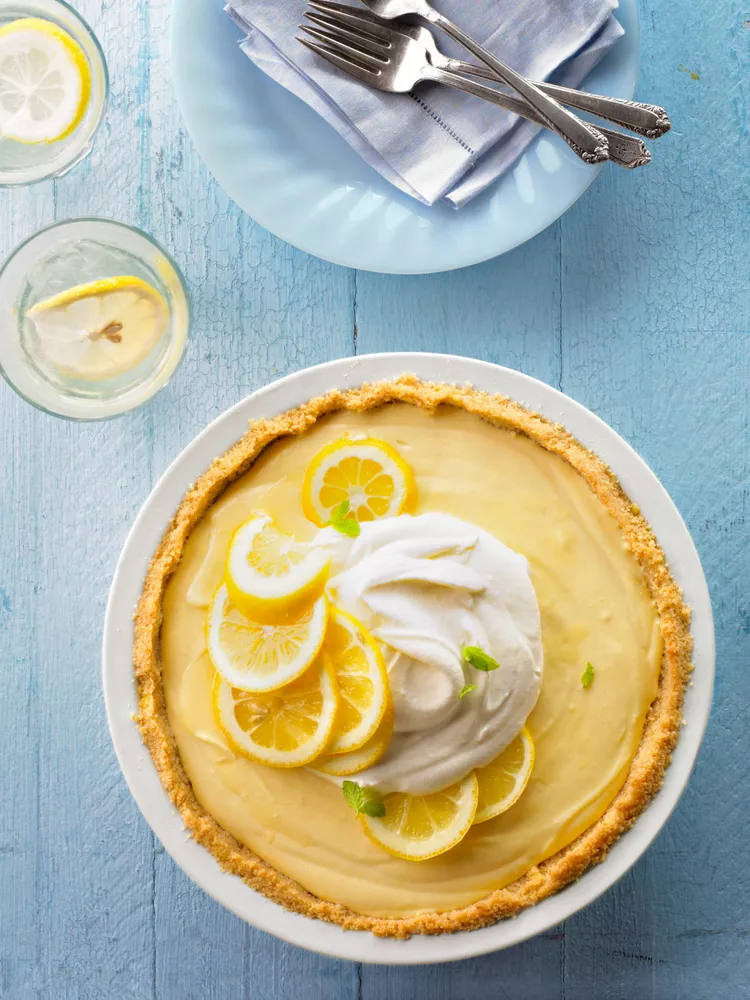 A lemon pie topped with whipped cream, lemon slices, and mint leaves, on a blue wooden table next to a plate with forks and two glasses of lemon water.