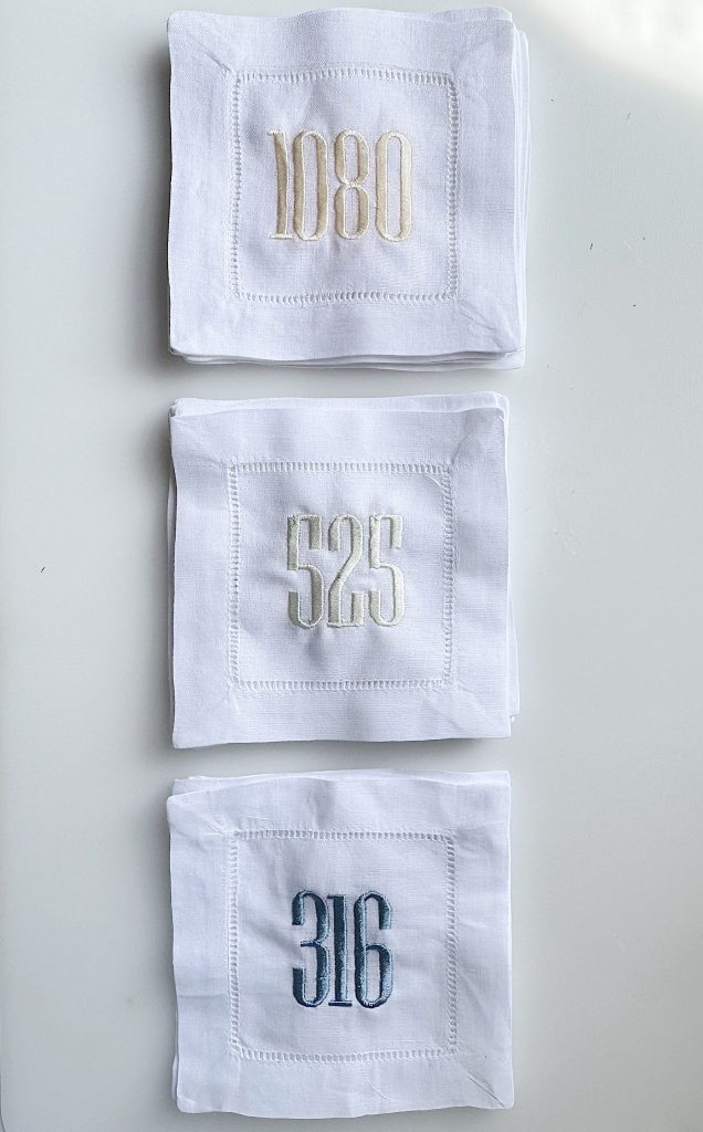 Three stacks of white cloth napkins, each embroidered with a different number: 1080 on top, 525 in the middle, and 316 on the bottom.