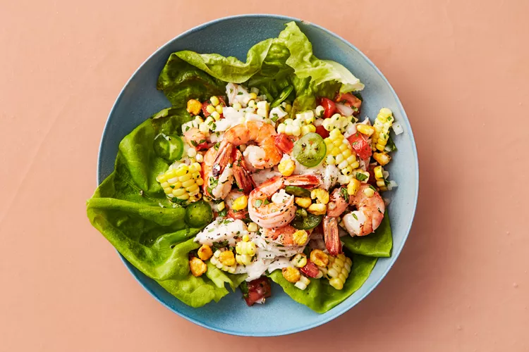 A blue plate with a salad of lettuce leaves, shrimp, corn, diced tomatoes, onions, jalapeño slices, and a creamy dressing on a peach-colored surface.