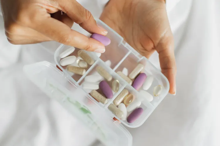 A person holds a pill organizer filled with various pills and capsules, picking up a purple oval tablet.