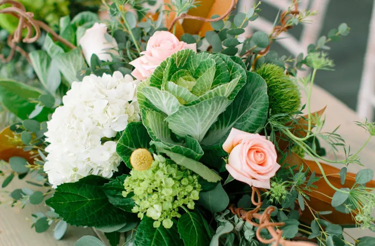 A floral arrangement featuring pink roses, white hydrangea, green leaves, cabbage, and assorted greenery.