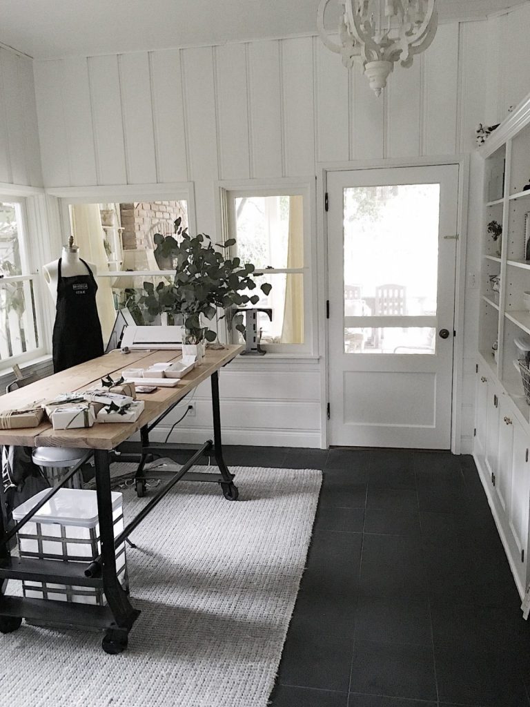 A bright, white-walled room with large windows, a worktable holding various items, a dress form, shelves on one side, and a door leading outside.