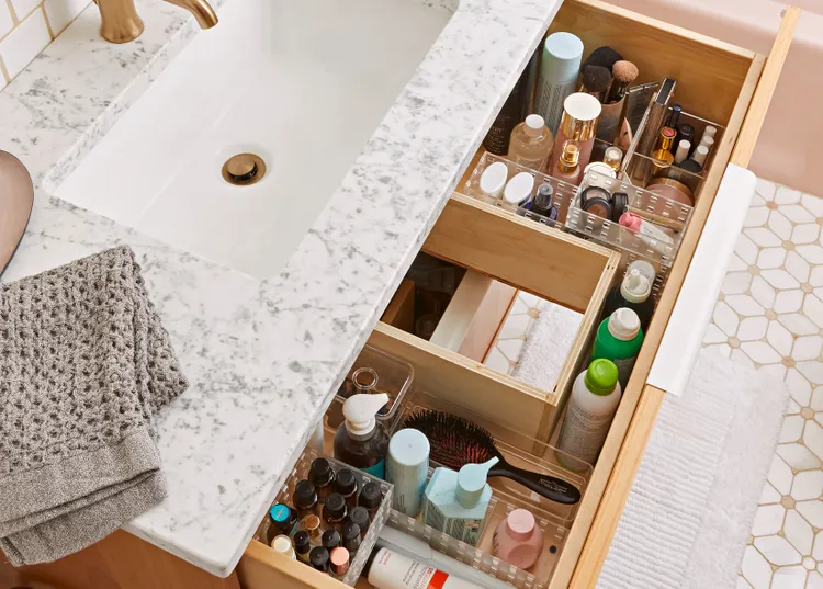Open bathroom drawer under a marble countertop sink, neatly organized with cosmetics, skincare bottles, towels, a hairbrush, and toiletries in clear and wooden dividers.