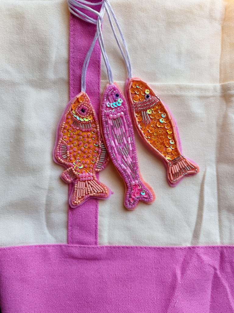 Three pink and orange fish-shaped ornaments with beads and sequins hang by thread against a cream and pink fabric background.