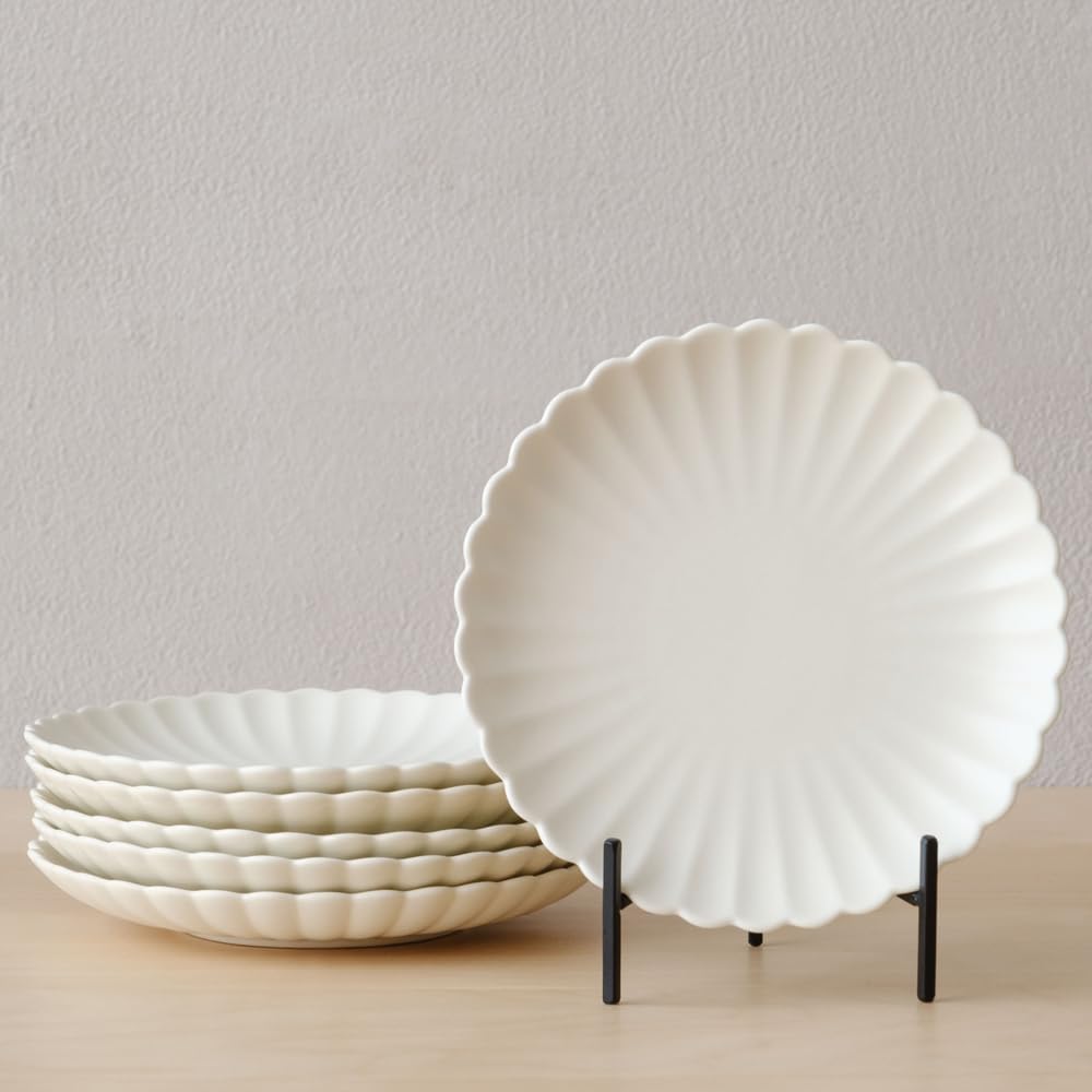 Six white, scalloped-edge ceramic plates are displayed; five are stacked on the left, and one stands upright on a black plate holder on a wooden surface against a plain wall—perfect for serving a vibrant grilled peach salad at your next gathering.
